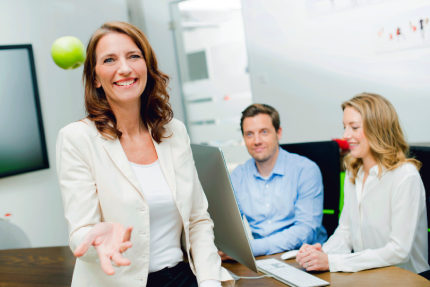 Betriebliche Gesundheitsmanagerin lacht in die Kamera, wirft einen grünen Apfel hoch. Im Hintergrund sitzen eine Kollegin und ein Kollege vor einem PC.