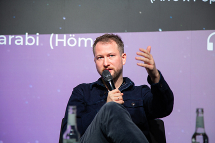 Rouven Bönisch sitzt auf einer Bühne und spricht in ein Mikro, das er in der Hand hält.